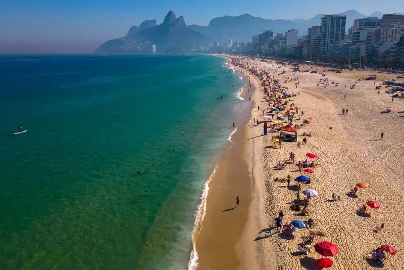 شاطئ مزدحم مع أشخاص يستمتعون بالشمس والبحر، وأشجار ونخيل على طول الساحل، مما يعكس أجواء الصيف المثالية للسفر.