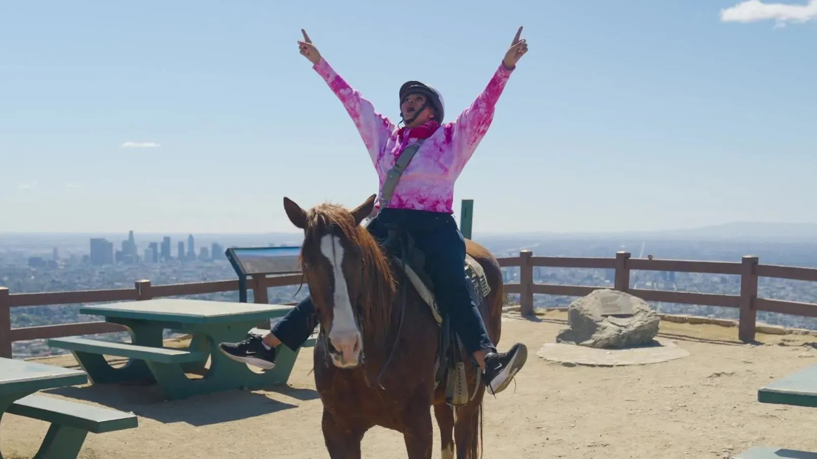 راكب على حصان في قمة غريفيث بارك، يرفع ذراعيه احتفالاً، مع إطلالة على المدينة خلفه. الحديقة تُظهر جمالها الطبيعي.