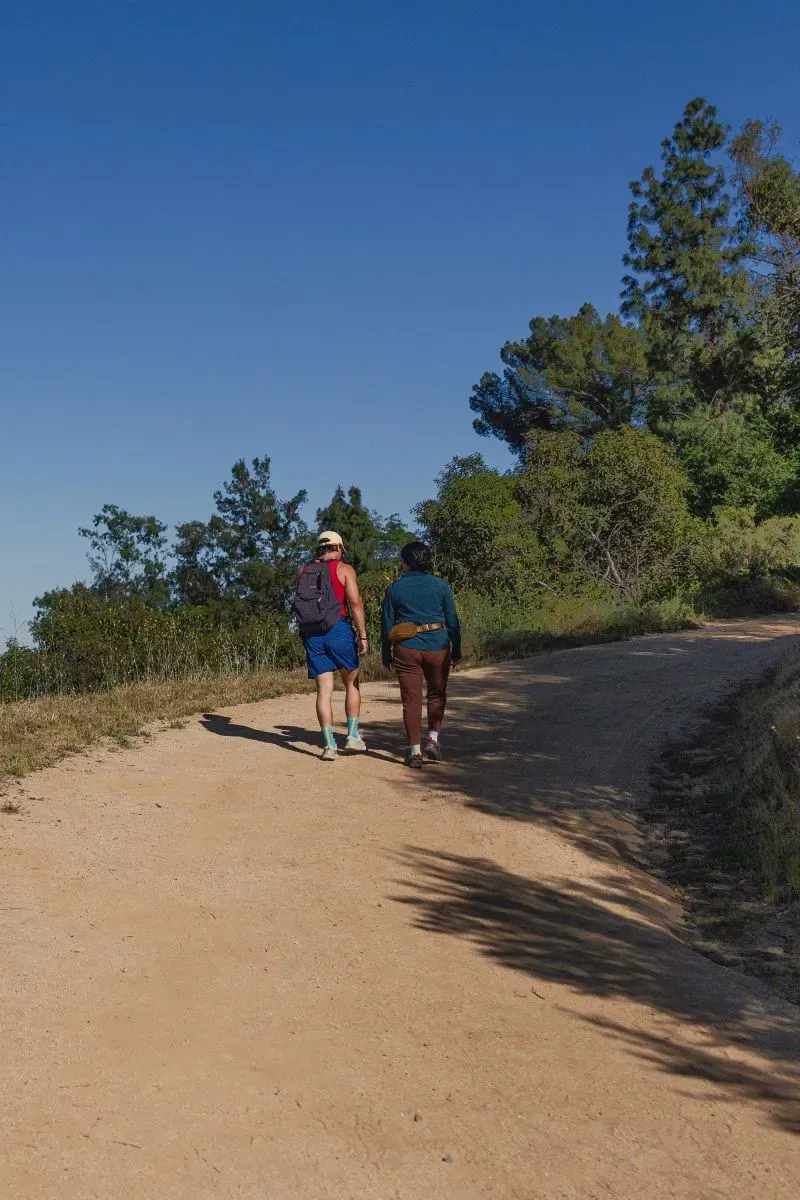 رجلان يسيران في مسار ترابي داخل حديقة غريفيث بارك، محاطين بأشجار خضراء تحت سماء زرقاء صافية، مما يعكس جمال الطبيعة في لوس أنجلوس.