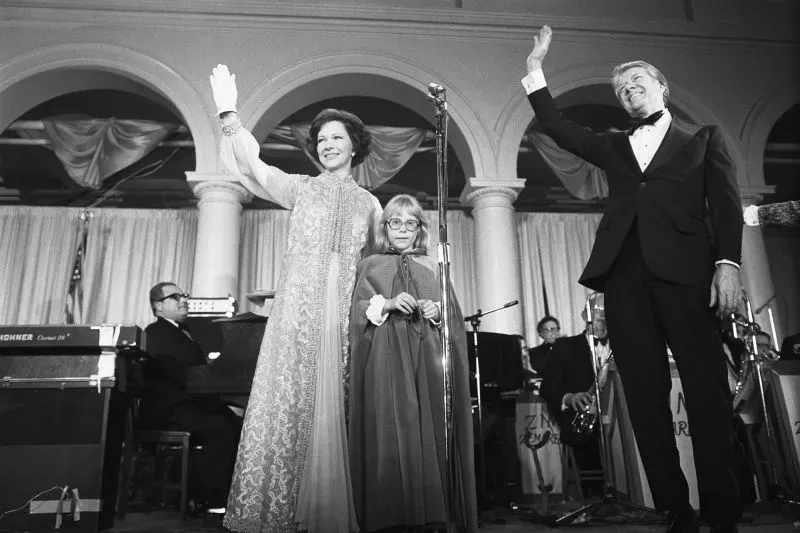 احتفال بمناسبة تنصيب الرؤساء حيث تظهر السيدة الأولى، ترتدي فستاناً مميزاً وتلوح للجمهور، مع خلفية موسيقية وعروض أخرى.