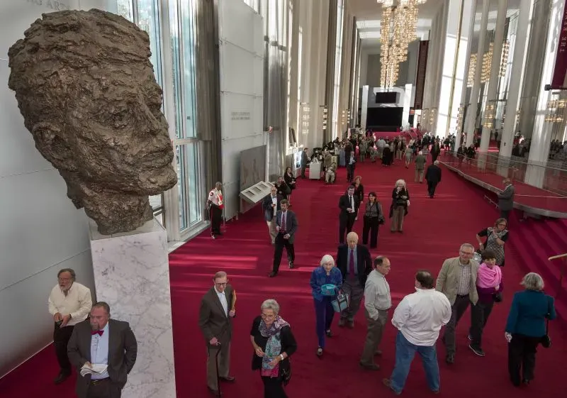 تمثال نصفي لجون كينيدي في مركز كينيدي للفنون، مع زوار يتجولون في قاعة واسعة ذات تصميم عصري وإضاءة مميزة.