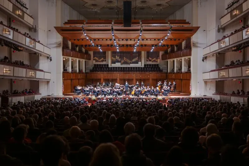 قاعة مركز كينيدي للفنون المسرحية مليئة بالجمهور أثناء عرض موسيقي، مع فرقة موسيقية على خشبة المسرح.