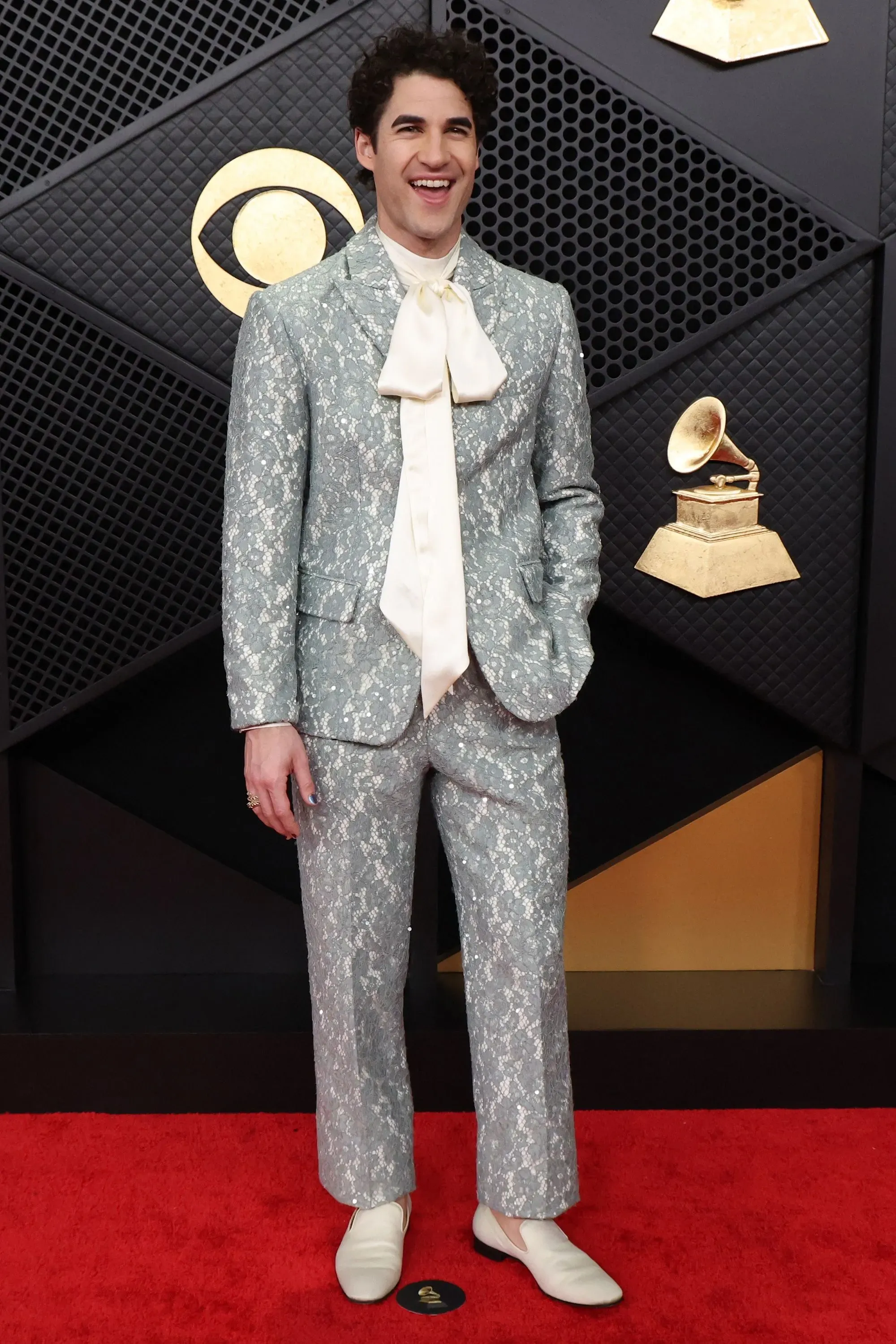 شخص يرتدي بدلة فضية مزخرفة مع ربطة عنق كبيرة، مبتسمًا على السجادة الحمراء في حفل توزيع جوائز غرامي، مع خلفية تتضمن شعار الجائزة.