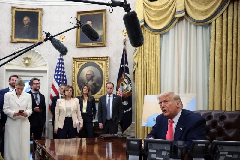 اجتماع في المكتب البيضاوي حيث يتحدث دونالد ترامب، محاطًا بمساعديه، مع ميكروفونات معلقة في الهواء، بينما تظهر صور تاريخية على الجدران.