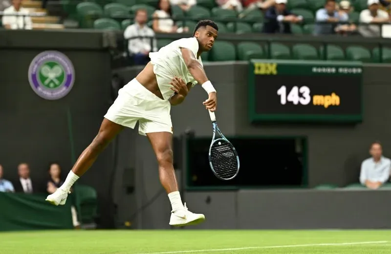 لاعب تنس يرسل الكرة بقوة في بطولة ويمبلدون، تظهر السرعة 143 ميلاً في الساعة على الشاشة خلفه، مع جمهور في الخلفية.