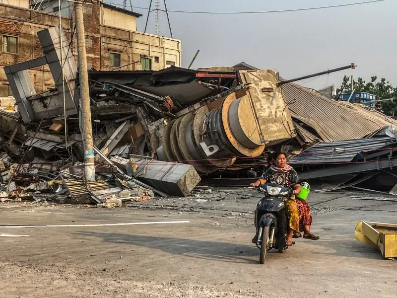 امرأة تقود دراجة نارية بجانب مباني منهارة في ساغاينغ، ميانمار، بعد الزلزال المدمر الذي خلف دماراً واسعاً ومأساة إنسانية.