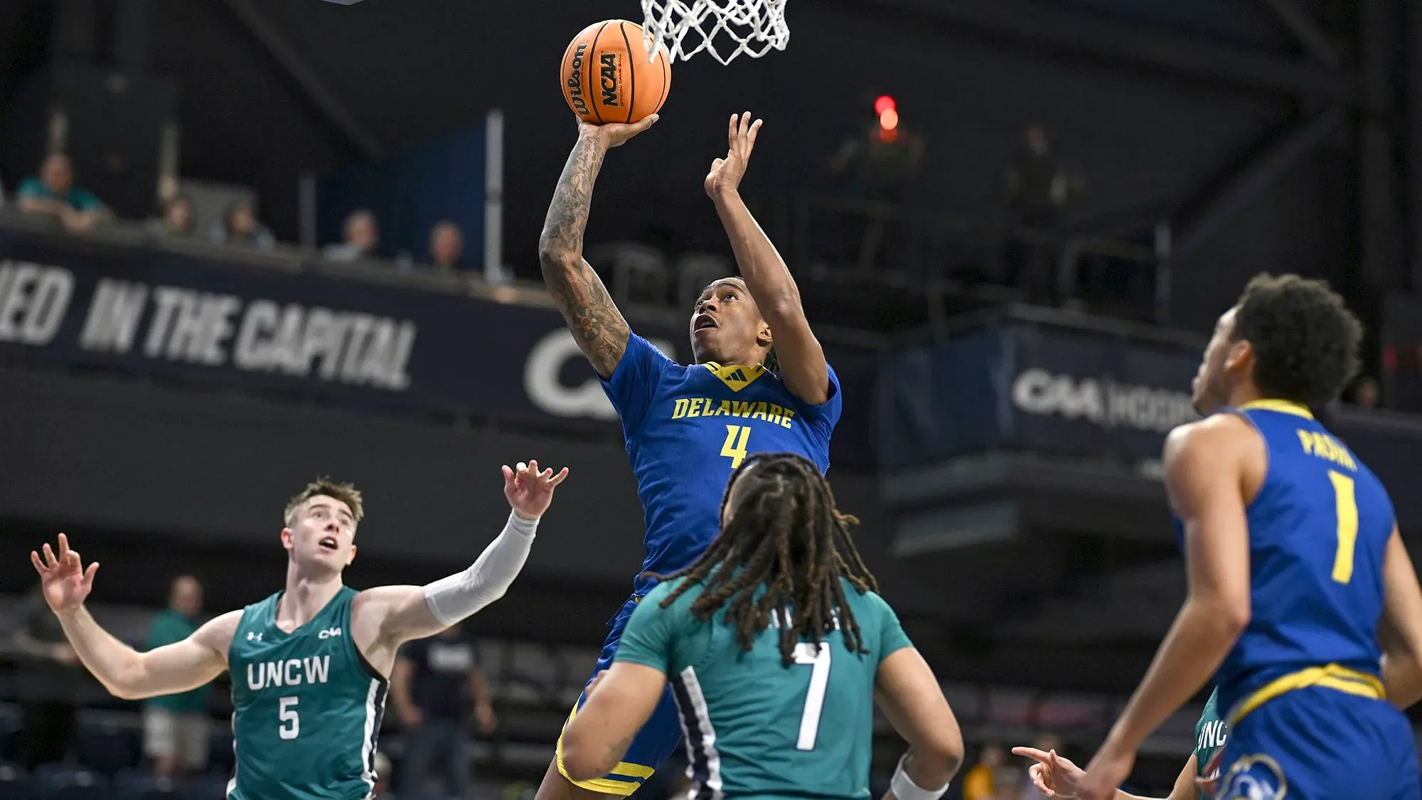 لاعب من جامعة ديلاوير يسجل نقطة خلال مباراة كرة السلة ضد جامعة نورث كارولينا ويلمنجتون، مع وجود لاعبين آخرين في الخلفية.