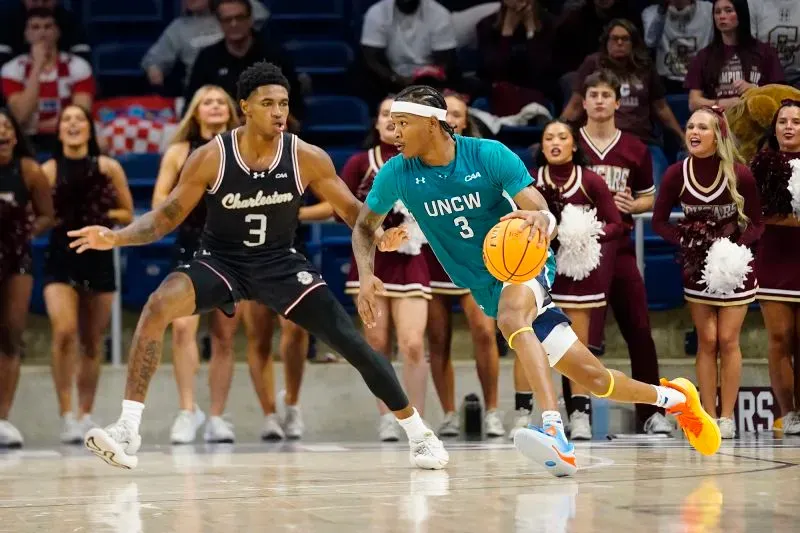 لاعب من جامعة نورث كارولينا ويلمنجتون يتجاوز مدافعًا من تشارلستون خلال مباراة كرة سلة في بطولة CAA، مع تشجيع حماسي من الجمهور.