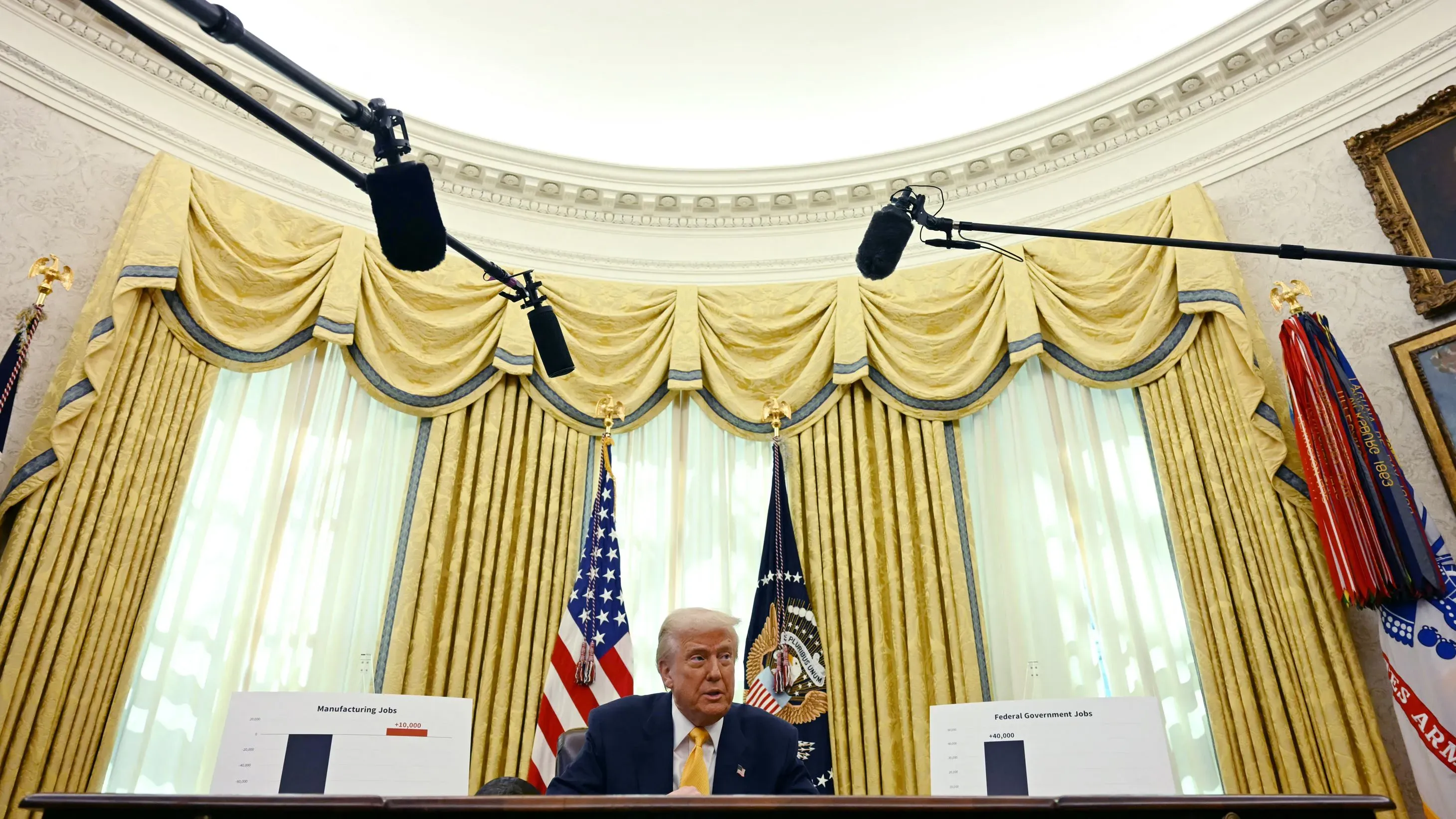 دونالد ترامب يجلس في المكتب البيضاوي، محاطًا بالستائر الذهبية، ويتحدث عن الاقتصاد مع وجود ميكروفونات أمامه.