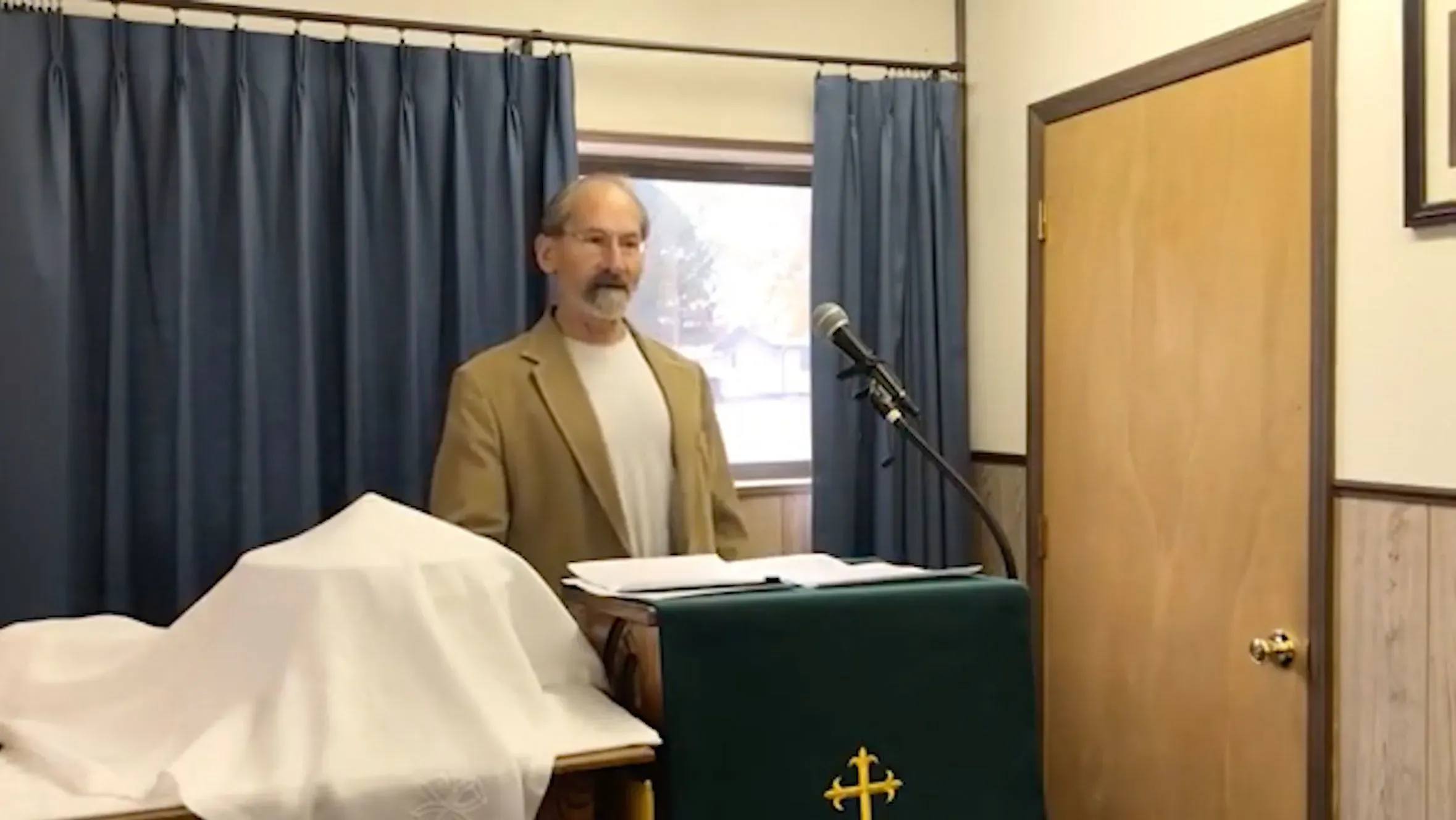 قس يتحدث في تجمع محلي، يرتدي بدلة، مع وجود ميكروفون أمامه وغطاء طاولة في الخلفية. تتعلق الصورة بانتخابات مجلس الشيوخ في نبراسكا.