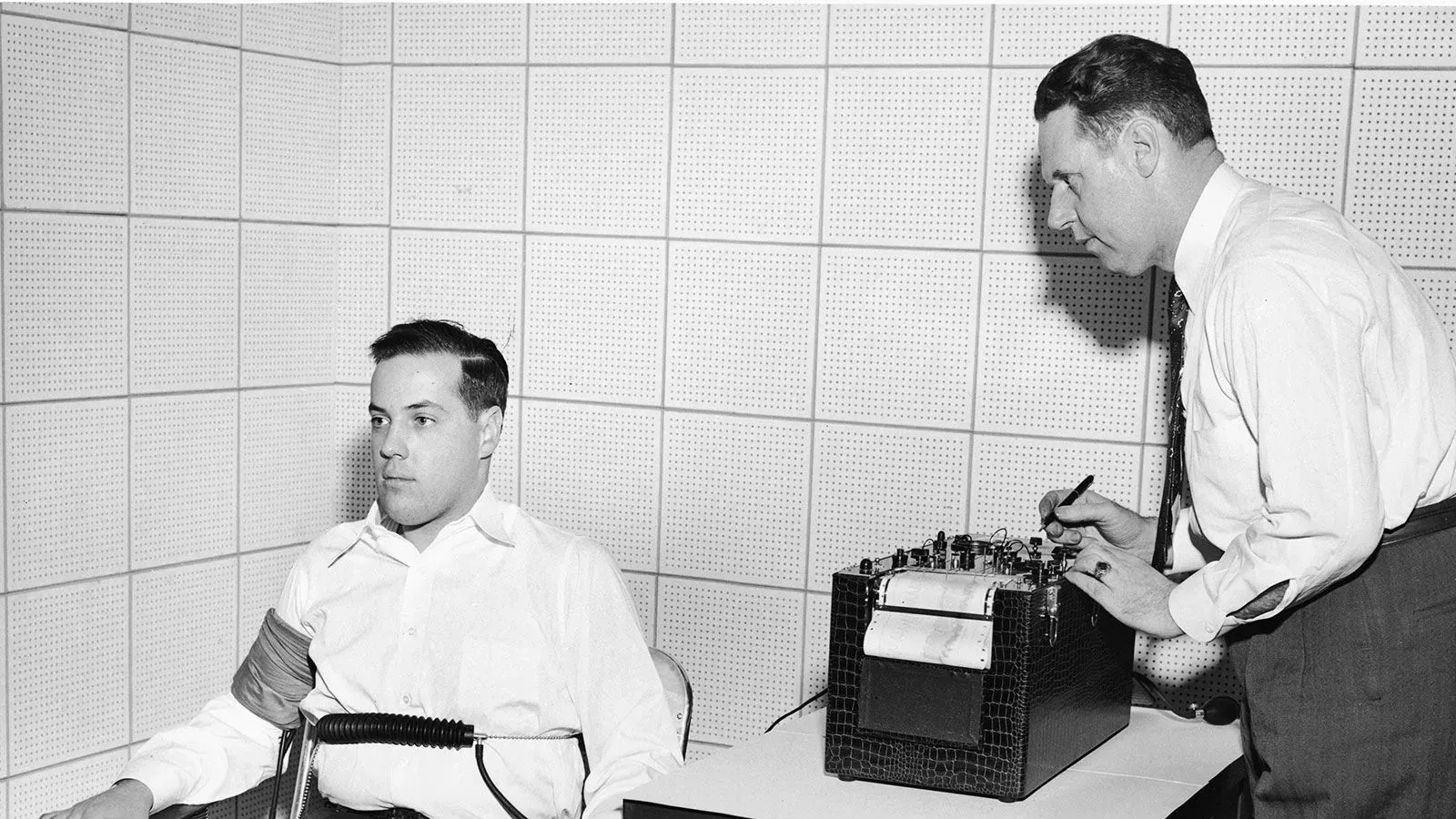 رجل يخضع لاختبار كشف الكذب في غرفة معزولة، بينما يقوم فاحص بتدوين الملاحظات على جهاز الكشف، مما يعكس التوتر في سياق الأمن القومي.