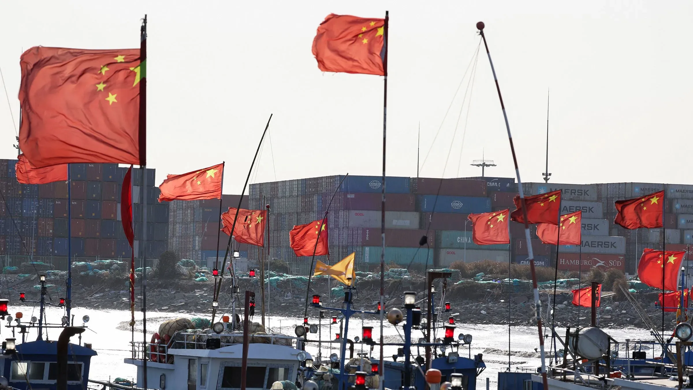 أعلام الصين ترفرف فوق مراكب صيد بالقرب من حاويات الشحن، مما يرمز إلى التوترات التجارية المتزايدة بين الولايات المتحدة والصين.