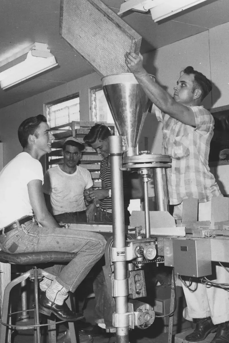 صورة تاريخية لرجلين يعملان في مصنع، حيث يقوم أحدهم بإدخال مادة في آلة بينما يجلس الآخر على مقعد.