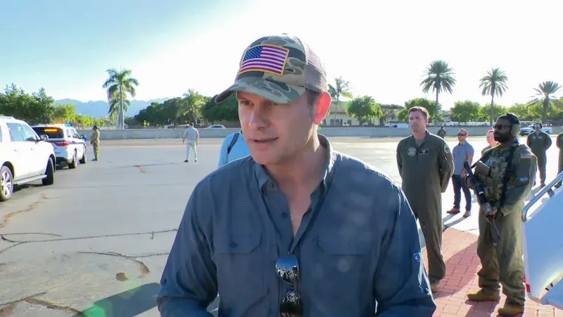 رجل يرتدي قبعة تحمل علم الولايات المتحدة، يتحدث مع مجموعة من العسكريين في خلفية تضم أشجار النخيل والمركبات، في سياق حديث عن الأمن القومي.