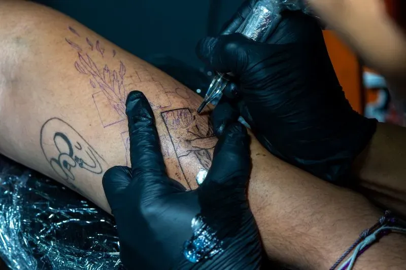 تظهر الصورة شخصًا يتلقى وشمًا على ذراعه، مع التركيز على تفاصيل التصميم، مما يعكس ثقافة الأوشام المرتبطة بالعصابات في السلفادور.