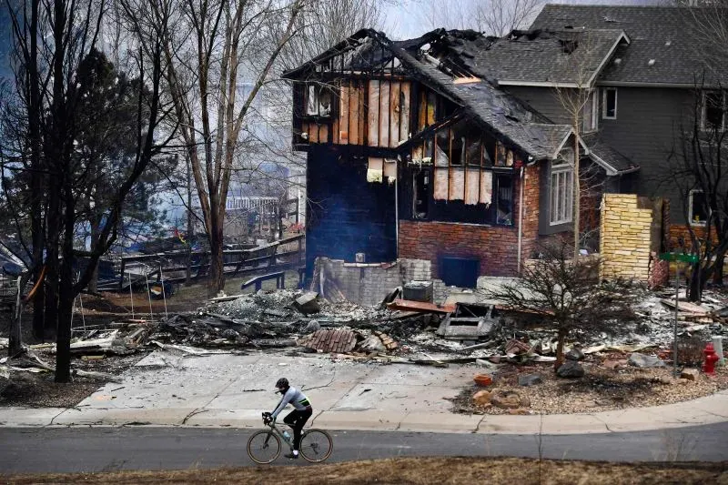 مشهد لبيت محترق جزئيًا في حي سكني بعد حريق مارشال، مع شخص يقود دراجة في المقدمة، مما يعكس أثر الكارثة على المجتمع.