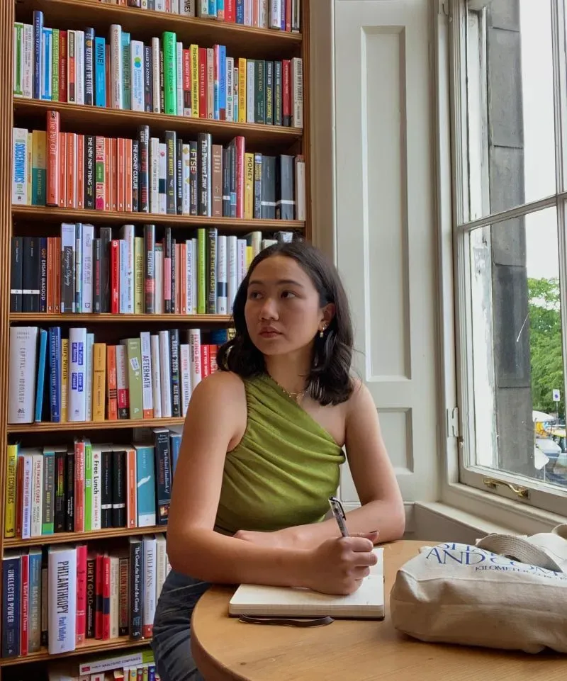 امرأة شابة تجلس في مكتبة، تكتب في دفتر ملاحظات، محاطة برفوف مليئة بالكتب الملونة. تعكس الصورة اهتمامها بالقراءة والتعلم.