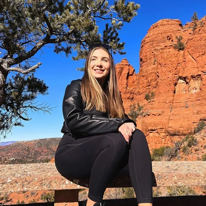 امرأة تبتسم وتجلس على حافة صخرية في المناظر الطبيعية الصحراوية، محاطة بأشجار خضراء وصخور حمراء تحت سماء زرقاء.