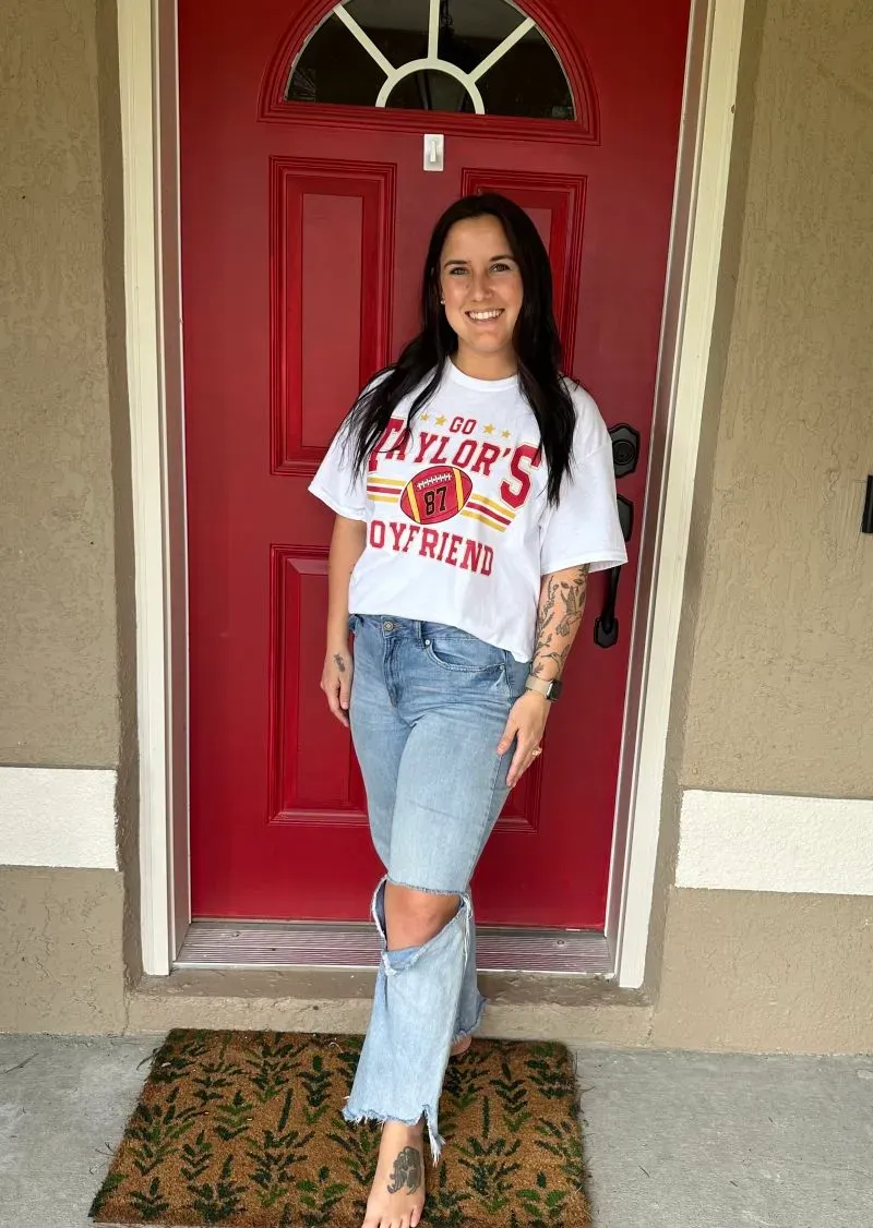 امرأة مبتسمة ترتدي قميصًا أبيض مع نقش وشورت جينز، تقف أمام باب منزلي أحمر، تعكس روح التحدي في مواجهة الاستهلاك المفرط.