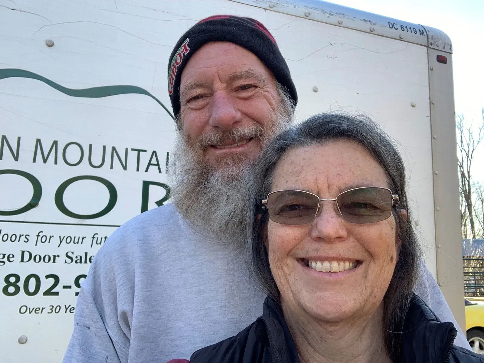 زوجان مبتسمان يقفان أمام شاحنة تحمل شعار شركة لتركيب الأبواب، يعكسان التحديات المالية المتعلقة بالتأمين الصحي.