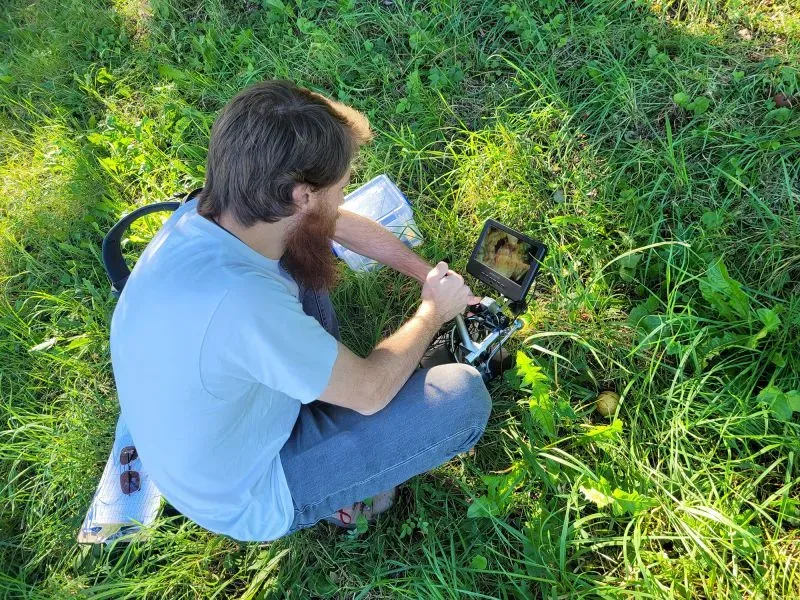 باحث يجلس على العشب ويستخدم جهاز عرض لمراقبة الديدان الخيطية أثناء دراستها في بيئة طبيعية.