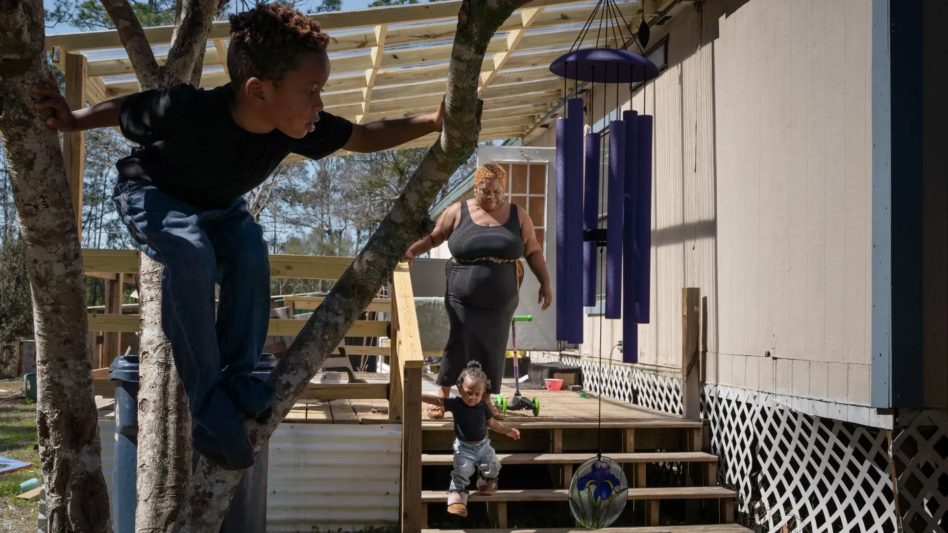 امرأة تسير على الدرج من منزل متنقل بينما يتسلق طفل شجرة، وآخر صغير يتبعها، مما يعكس الحياة الأسرية في فلوريدا.