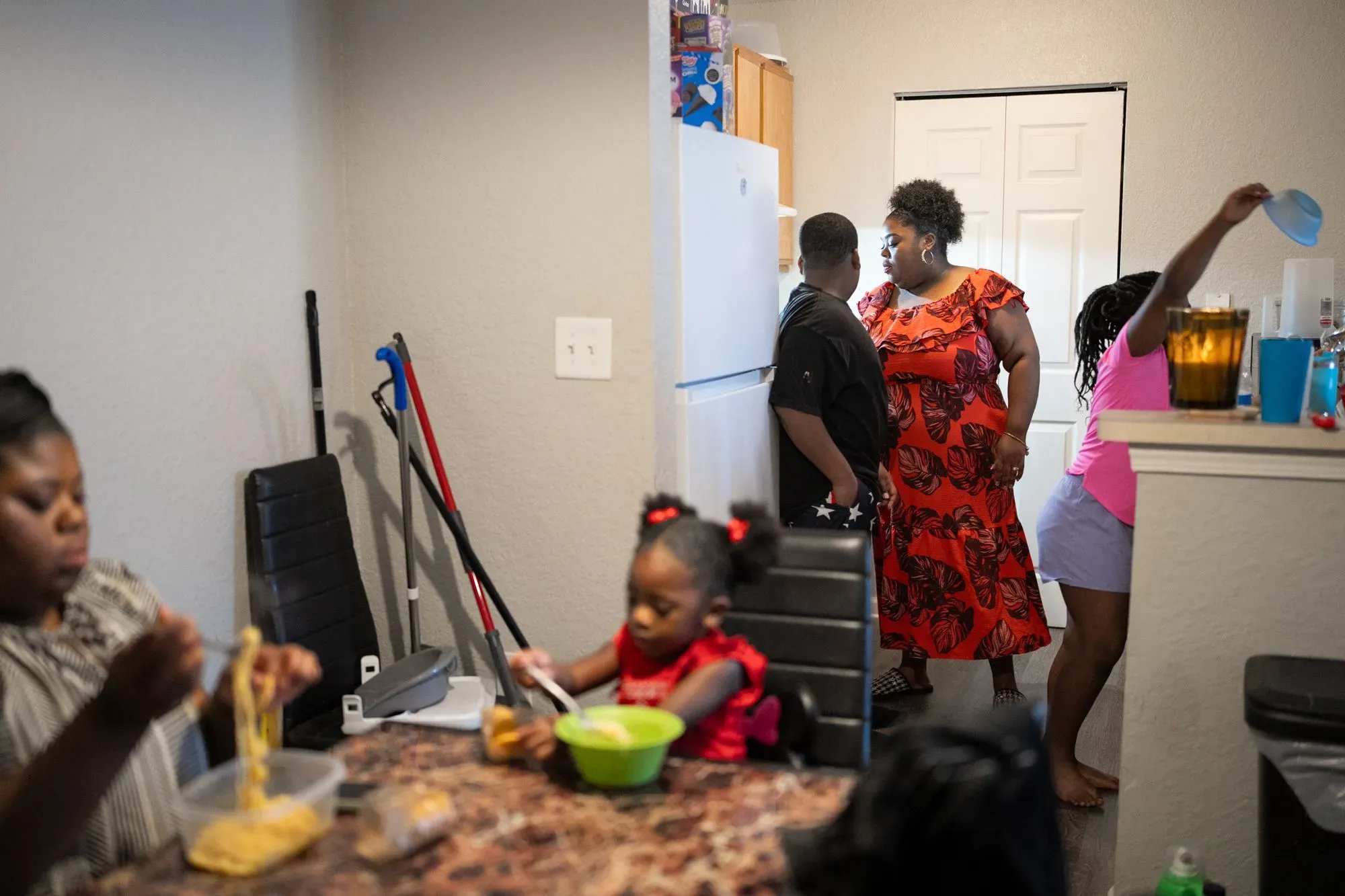 عائلة تتجمع في مطبخ بسيط، حيث تقوم امرأة بارتداء فستان مزخرف بإعداد الطعام، بينما الأطفال مشغولون بالأكل واللعب.