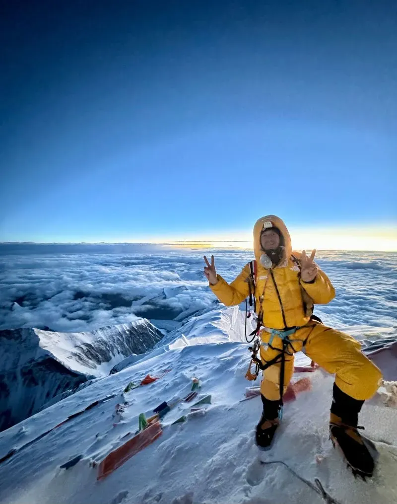 رجل يرتدي ملابس تسلق صفراء في قمة إيفرست، مبتسمًا ويعرض علامة النصر، مع منظر للغيوم في الخلفية.