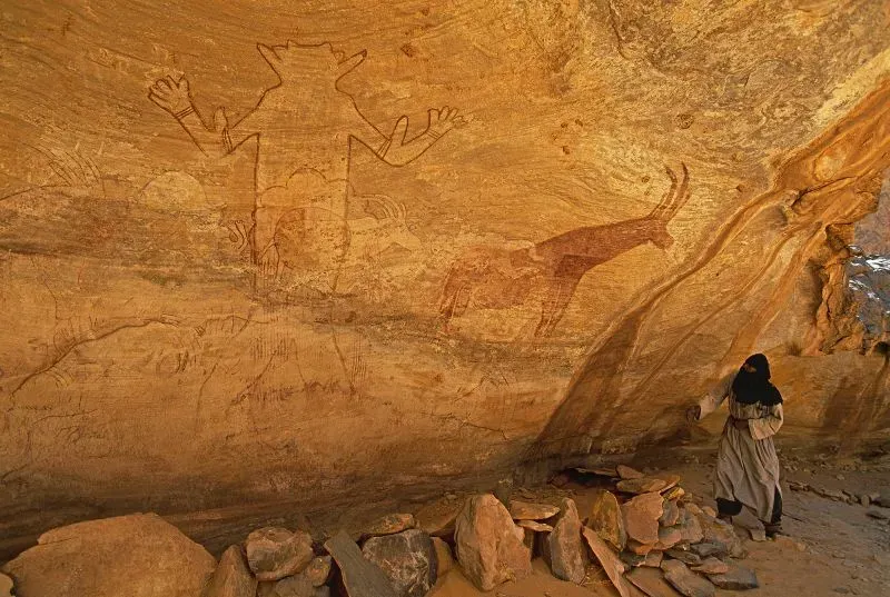 نقوش صخرية قديمة في منتزه الطاسيلي ناجر الوطني، مع سائح يقف أمامها، تعكس تاريخ الجزائر وثقافتها الغنية.