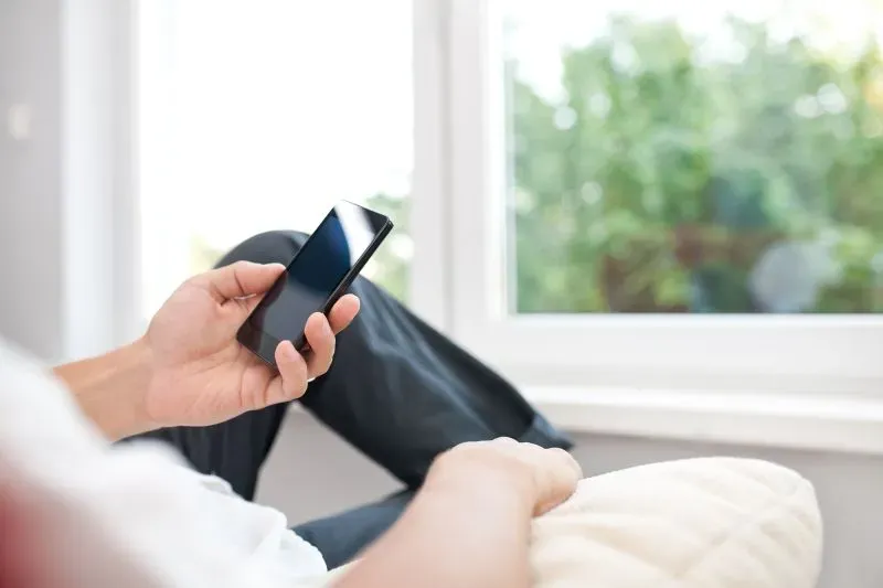 شخص يجلس على أريكة، ممسكًا بهاتفه الذكي، بجوار نافذة تطل على منظر طبيعي، مما يعكس لحظة استرخاء أو تشتت انتباه.