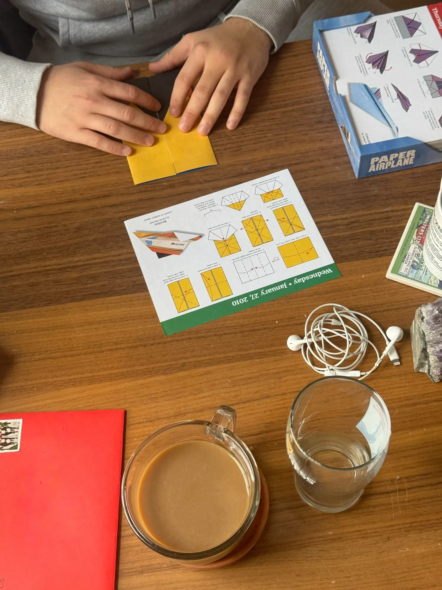 يد تظهر يدين تعملان على طي ورقة صفراء لصنع طائرة ورقية، مع وجود كوب من القهوة وزجاجة ماء ومخطط توجيهي على الطاولة.