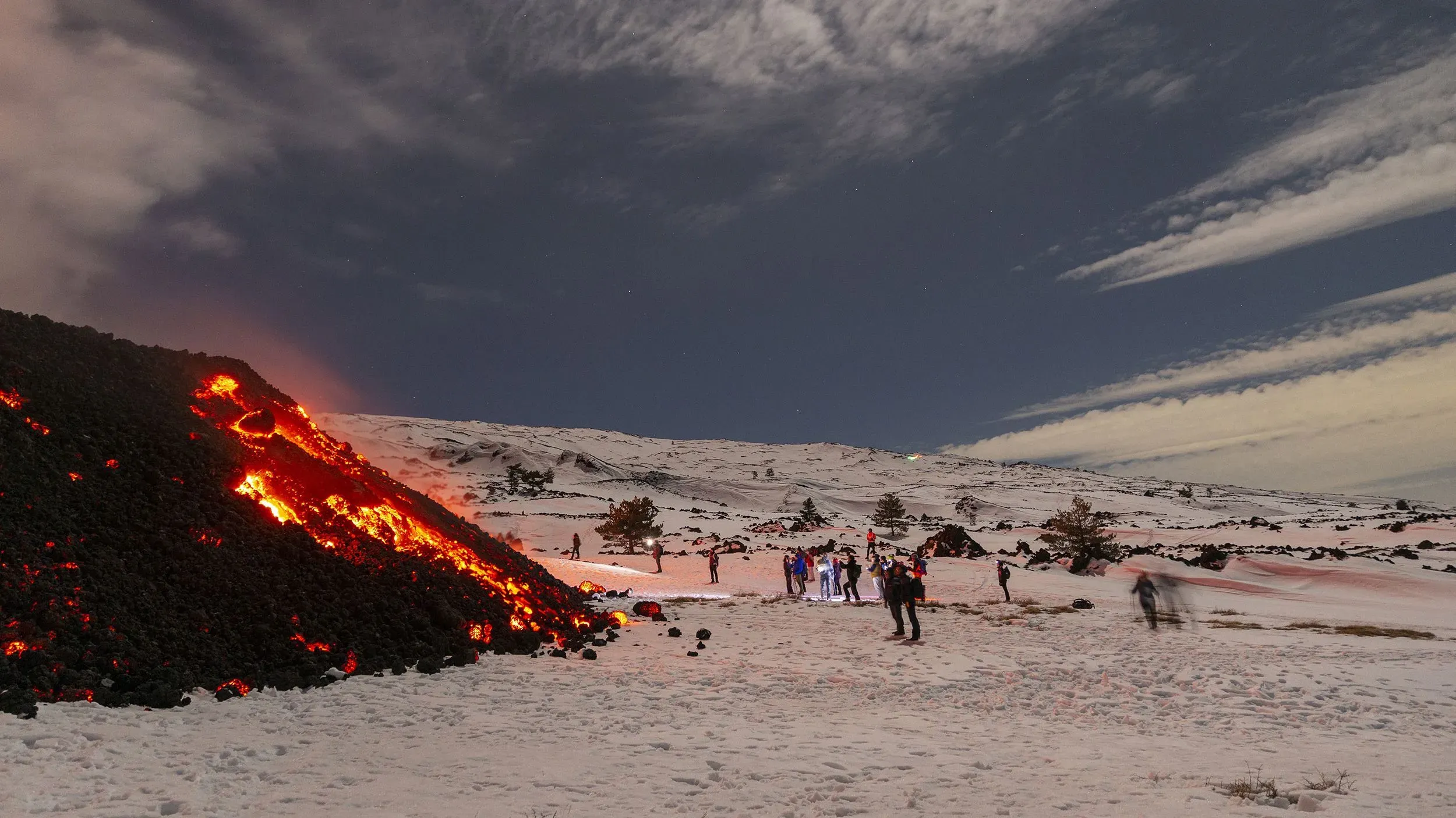 حمم بركانية تتدفق على جبل إتنا المغطى بالثلوج، مع مجموعة من المتفرجين في الخلفية، مما يبرز النشاط البركاني المذهل في صقلية.