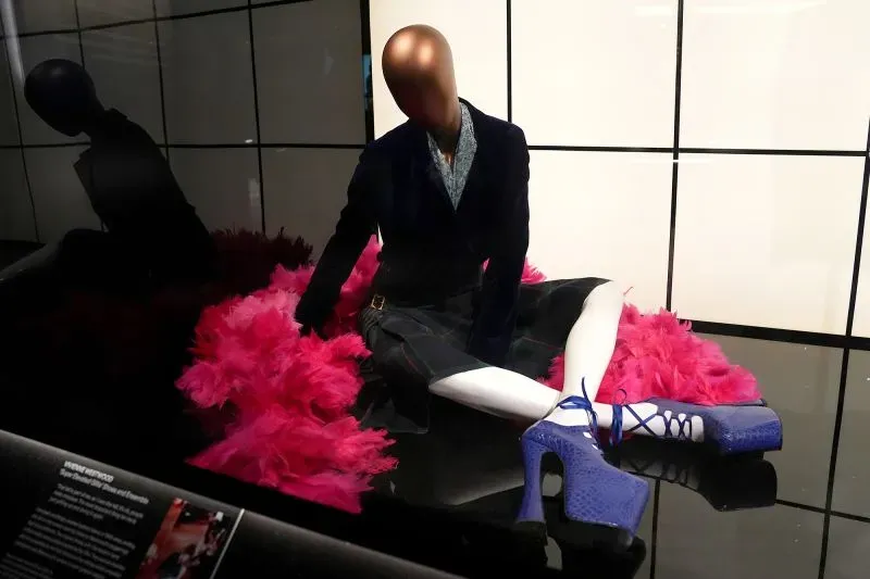 مجسم لعارضة أزياء ترتدي حذاءً عالي الكعب باللون الأزرق الفاتح، محاطاً بغطاء من الريش الوردي، يعكس لحظة تاريخية في عالم الموضة.