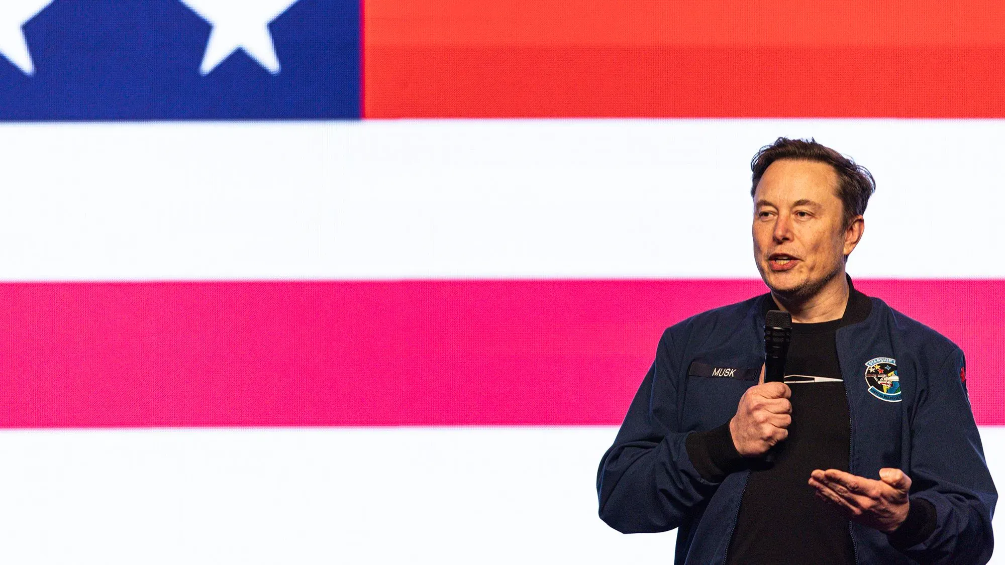 إيلون ماسك يتحدث خلال حدث، خلفه علم الولايات المتحدة، مع التركيز على قضايا التعريفات الجمركية وتأثيرها على تسلا.