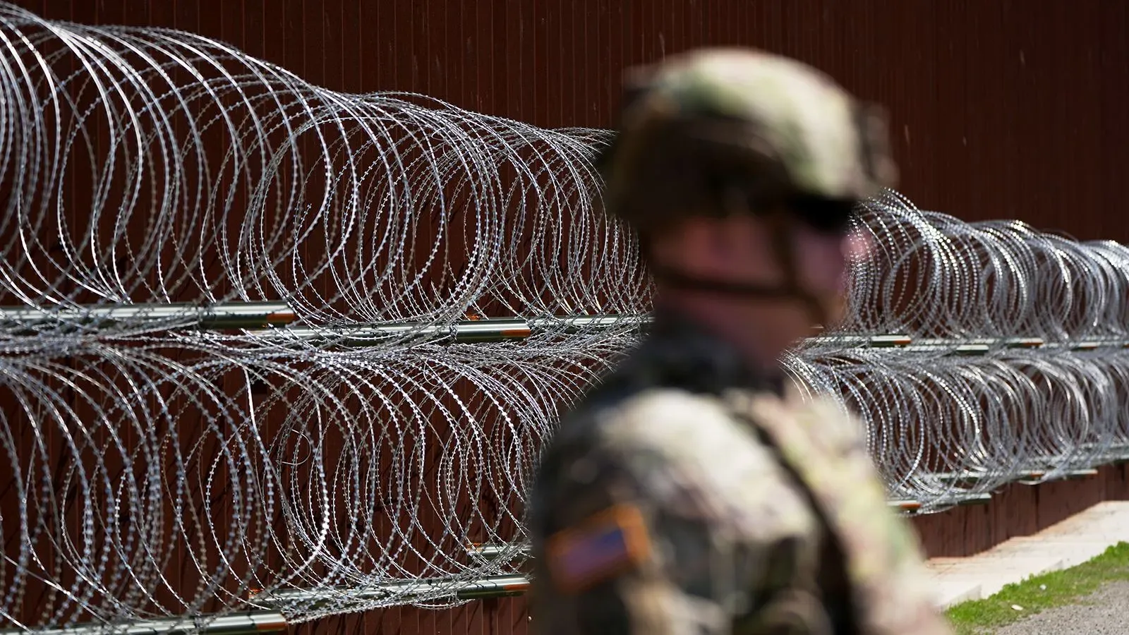 جندي يرتدي زيًا عسكريًا يقف أمام سياج شائك، مما يعكس الأوضاع الأمنية والمناقشات حول تصنيف المهاجرين كـ"مقاتلين أعداء".
