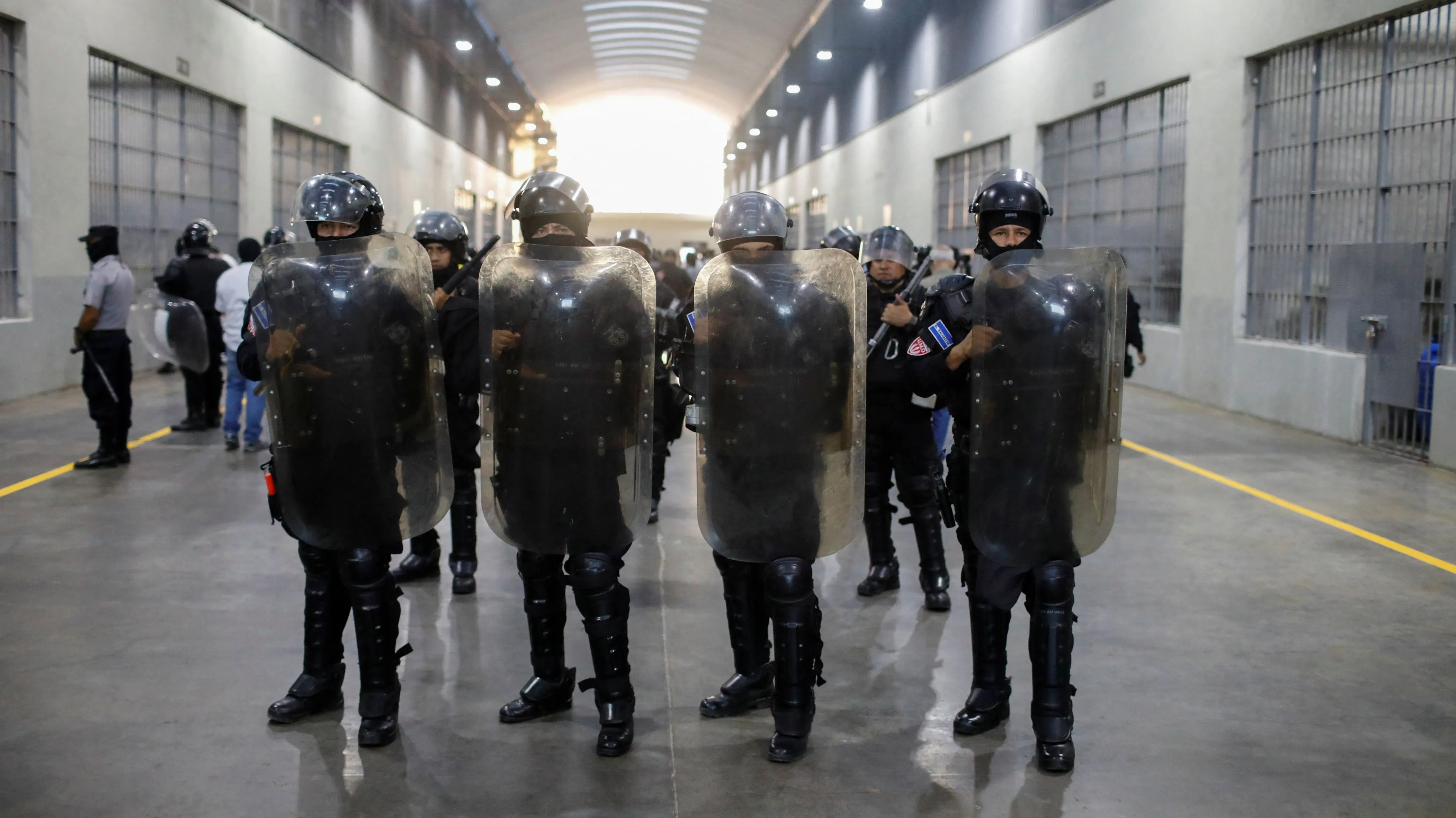 عناصر من الشرطة السلفادورية يرتدون دروعاً واقية، يقفون في ممر داخل سجن كبير، في سياق جهود إدارة ترامب لترحيل المهاجرين.