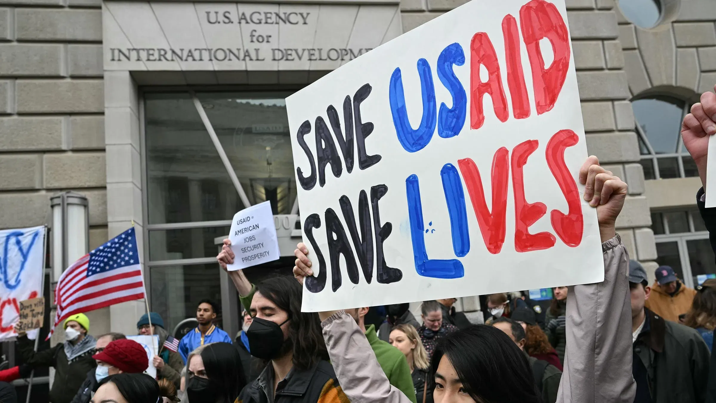 محتجون يحملون لافتة مكتوب عليها "أنقذوا الوكالة الأمريكية للتنمية الدولية، أنقذوا الأرواح" أمام مبنى الوكالة، مع العلم الأمريكي في الخلفية.
