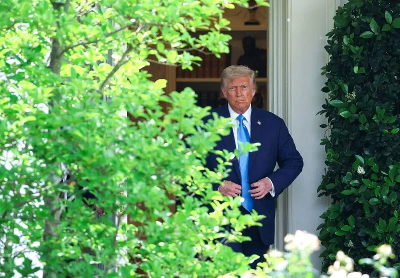 ترامب يخرج من البيت الأبيض محاطًا بالأشجار، مع تعبير جدي، في سياق تصاعد التوترات حول سياساته الاقتصادية.