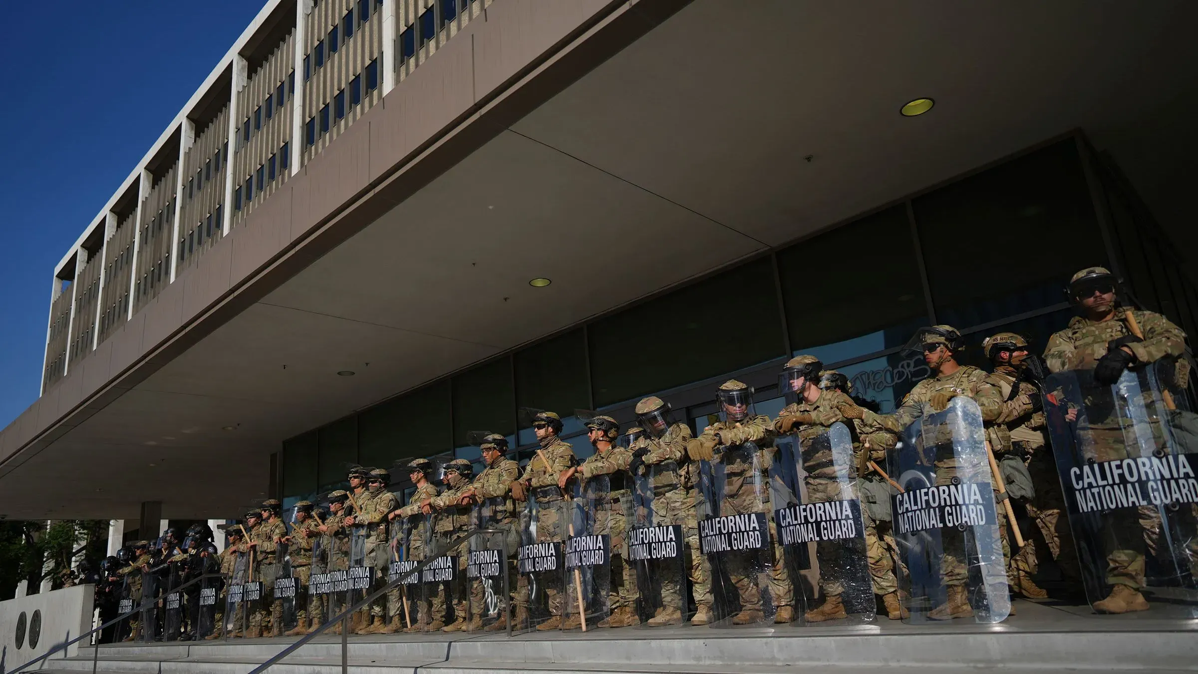 جنود من الحرس الوطني في كاليفورنيا يقفون في صفوف أمام مبنى حكومي، مستعدين لمواجهة الاحتجاجات المحتملة وتعزيز الأمن.
