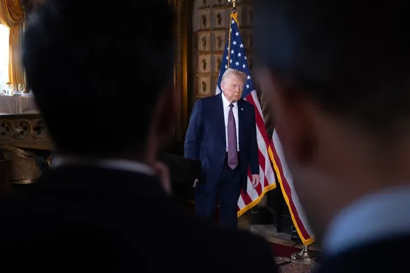 ترامب يقف أمام علم الولايات المتحدة، مع ظهور شخصين في الخلفية، في سياق مناقشات حول تأثير سوق السندات على السياسات الاقتصادية.