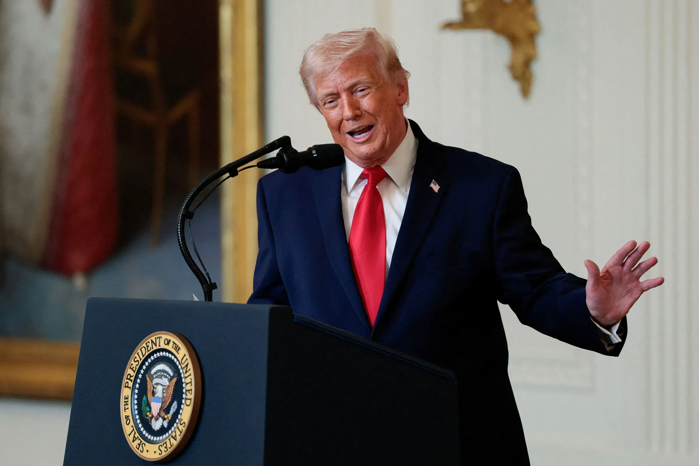 تظهر الصورة الرئيس الأمريكي السابق دونالد ترامب وهو يتحدث أمام ميكروفون، مرتديًا بدلة زرقاء وقميصًا أبيض مع ربطة عنق حمراء، في سياق مناقشة السياسة الأمريكية.