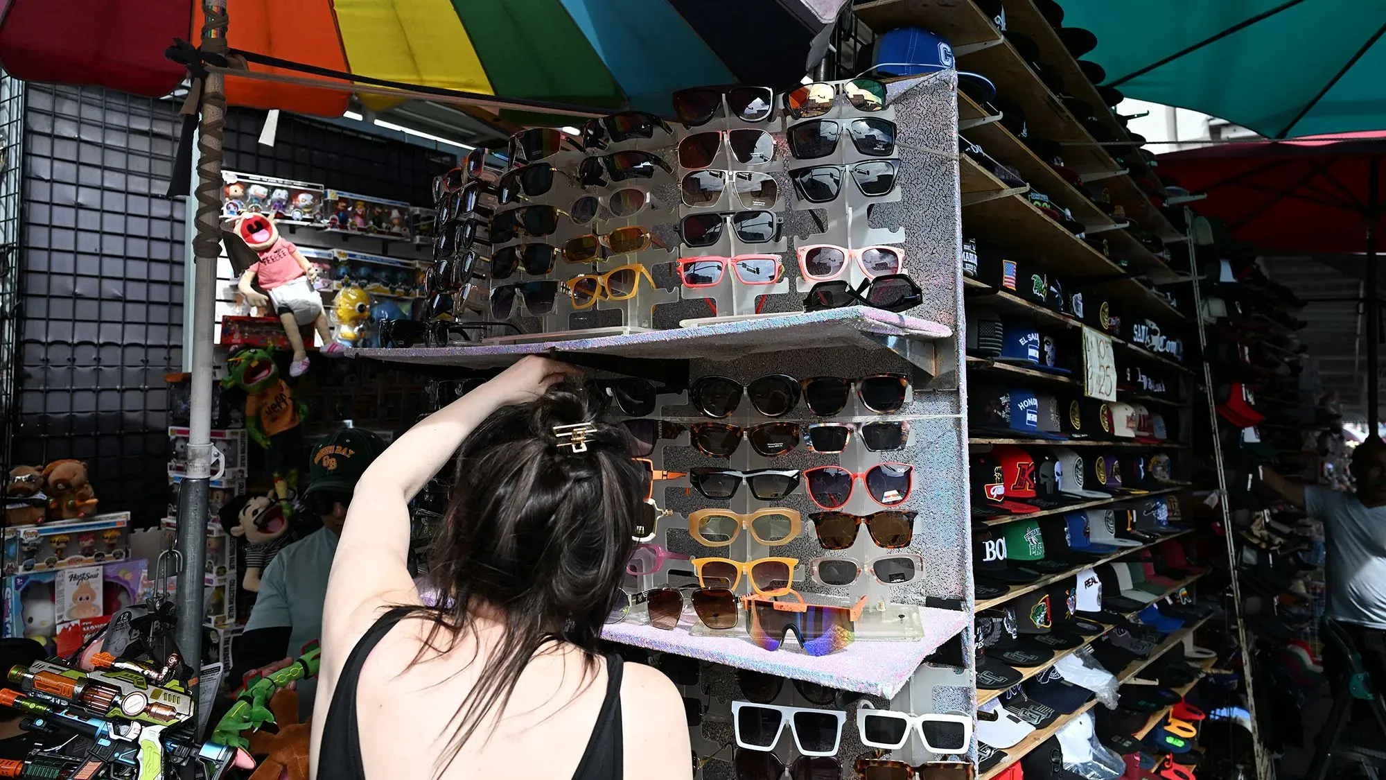 امرأة تفتش في رف من النظارات الشمسية في سوق، مع دمى وألعاب خلفها، تعكس تأثير الرسوم الجمركية على أسعار السلع.
