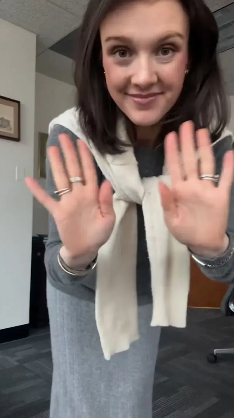 امرأة مبتسمة ترتدي ملابس أنيقة في مكتبها، تلوح بيديها، بينما تسجل فيديو لمشاركة أسلوبها على إنستغرام.