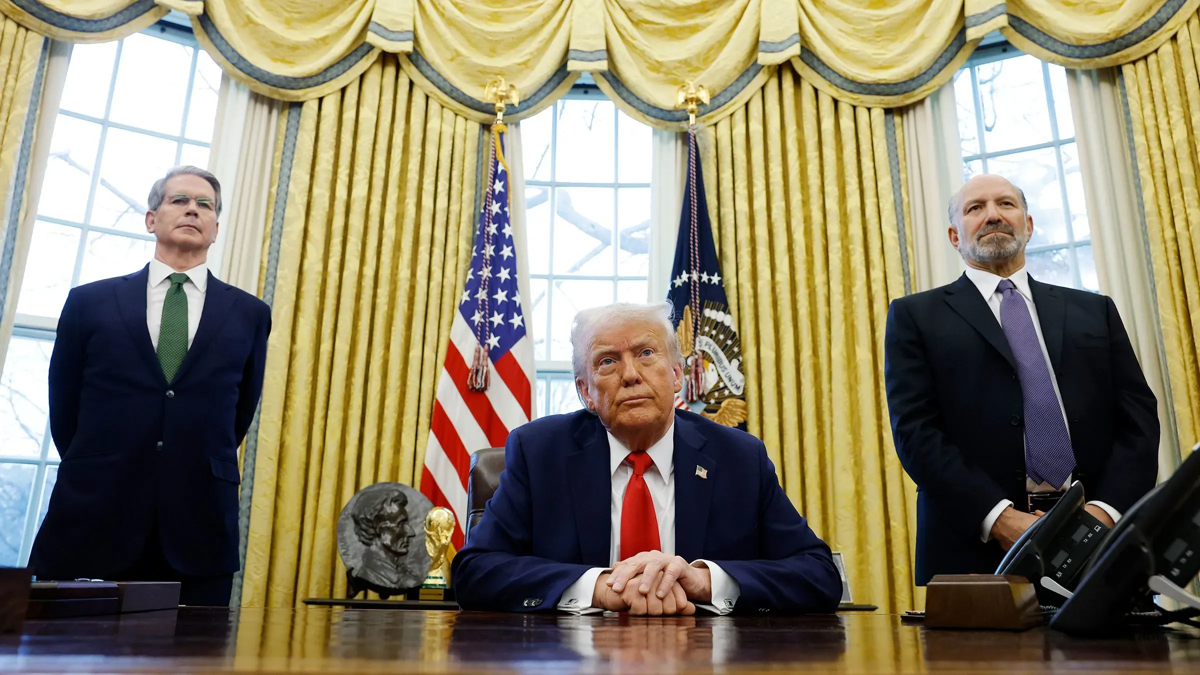 صورة لدونالد ترامب يجلس في المكتب البيضاوي، محاطًا بمستشاريه، مع العلم الأمريكي خلفه، في سياق مناقشة السياسات الاقتصادية.