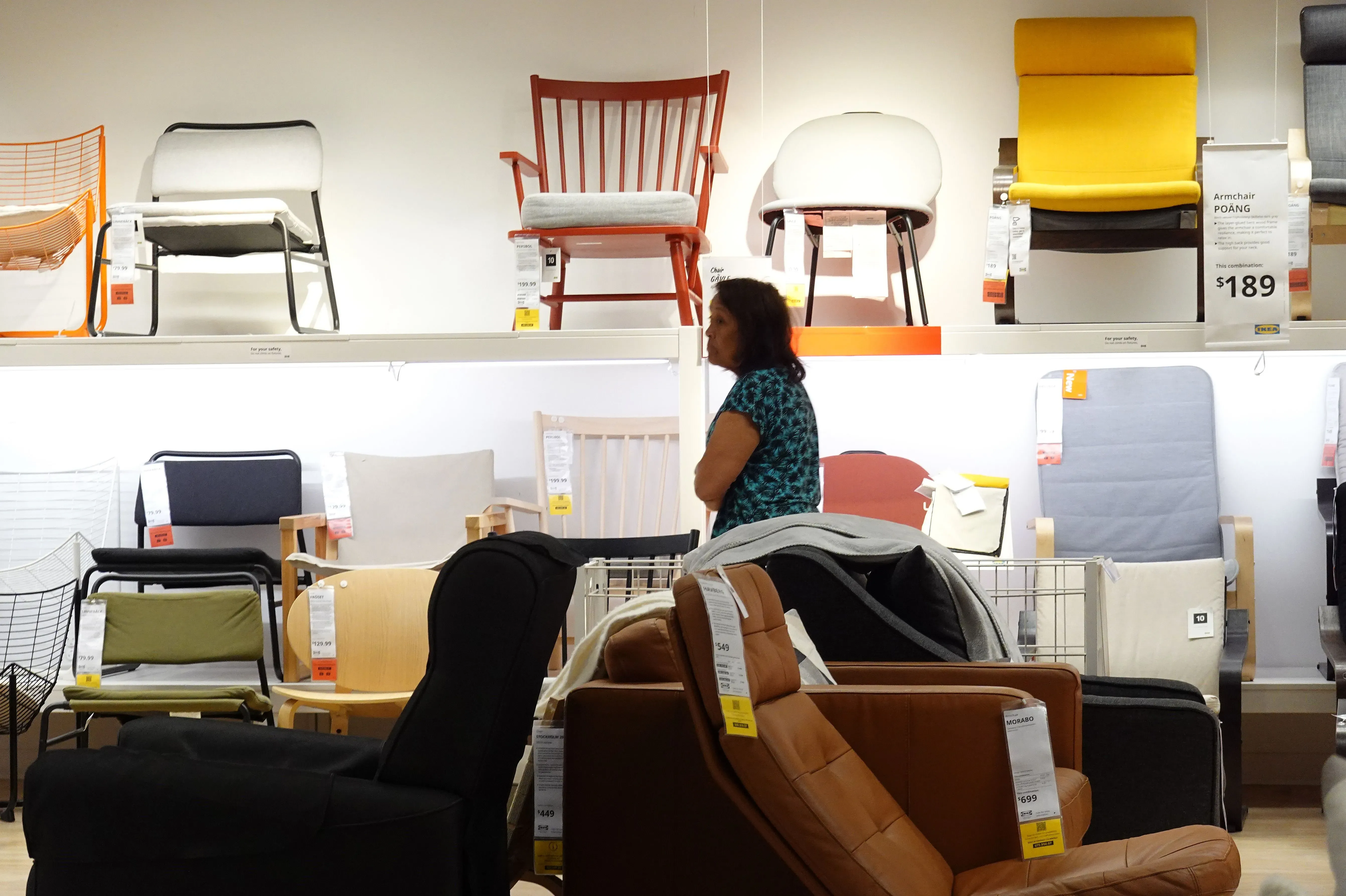 امرأة تتجول بين مجموعة من الكراسي والأثاث المنجد في متجر، حيث تظهر الأسعار والعلامات على المنتجات.