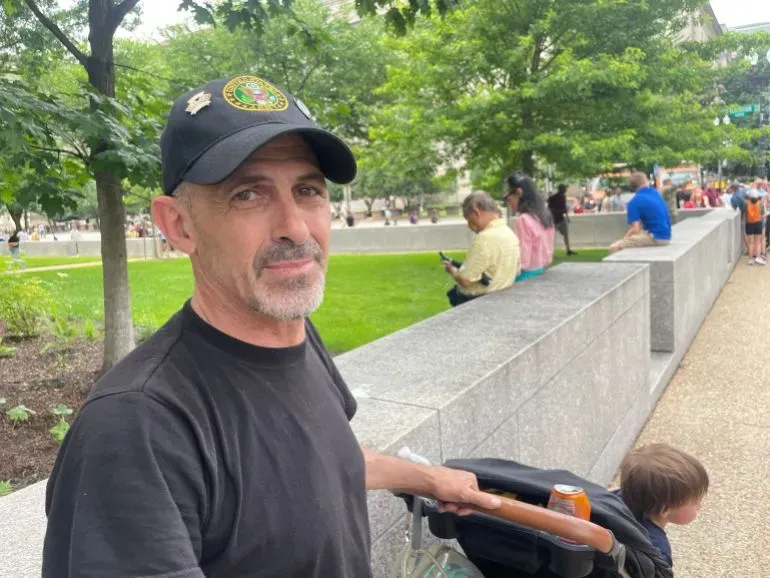 رجل مسن يرتدي قبعة تحمل شعارًا عسكريًا، يجلس في حديقة عامة وسط مجموعة من الناس، بينما يبدو طفل صغير بجانبه.