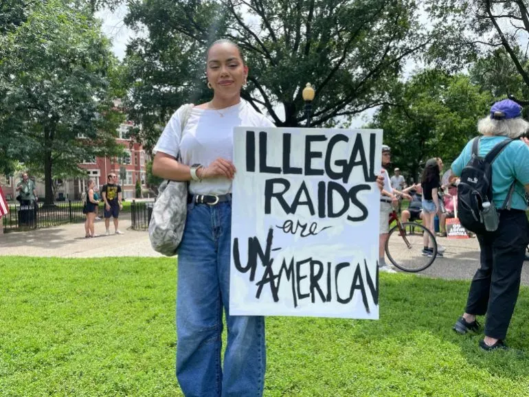 امرأة تحمل لافتة مكتوب عليها "المداهمات غير القانونية غير أمريكية" خلال احتجاج في واشنطن، مع وجود حشد في الخلفية.