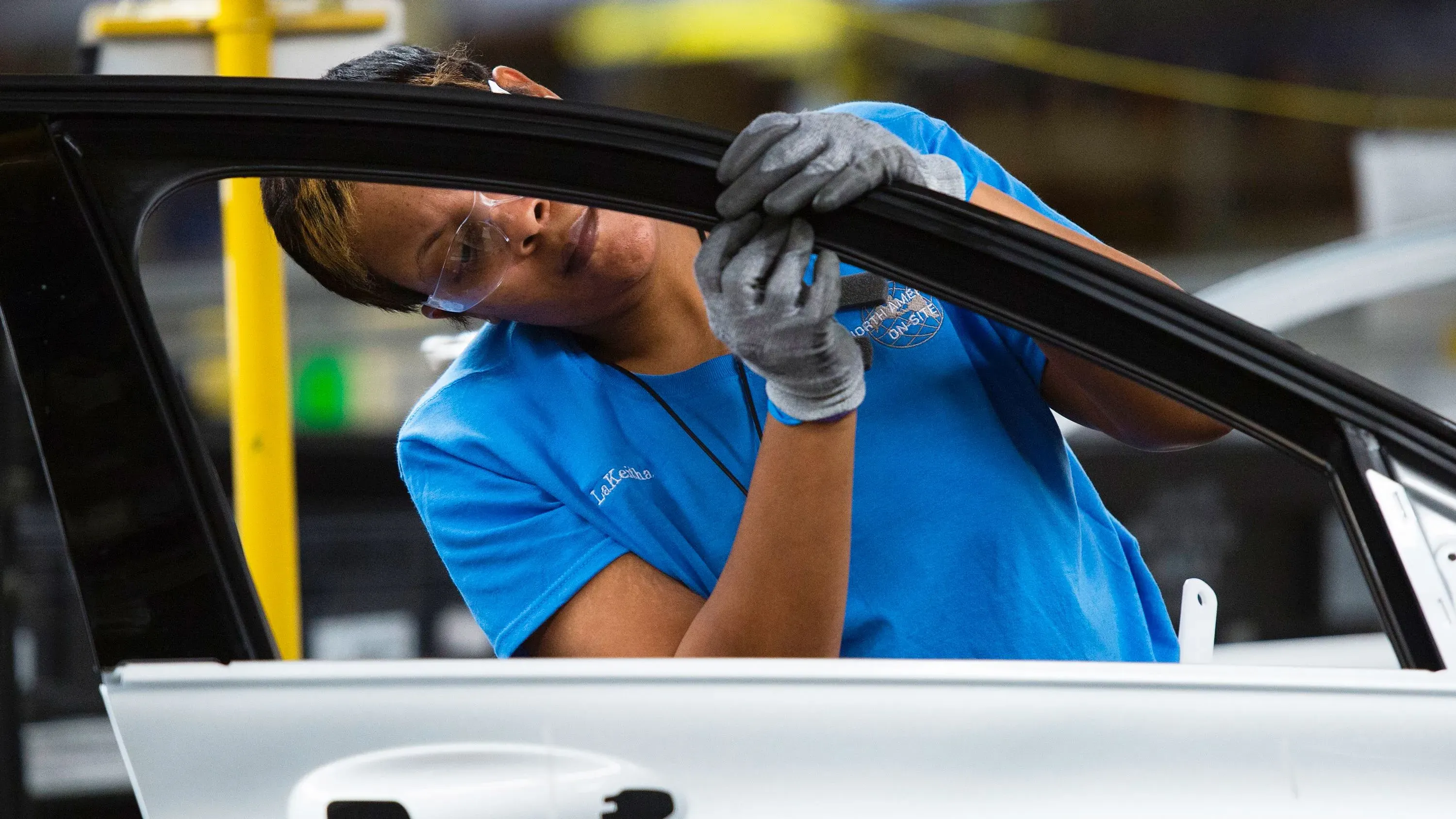 امرأة ترتدي نظارات وقبعة تعمل في مصنع سيارات، تركز على تركيب جزء من السيارة، مما يعكس تأثير الأتمتة على صناعة السيارات الأمريكية.