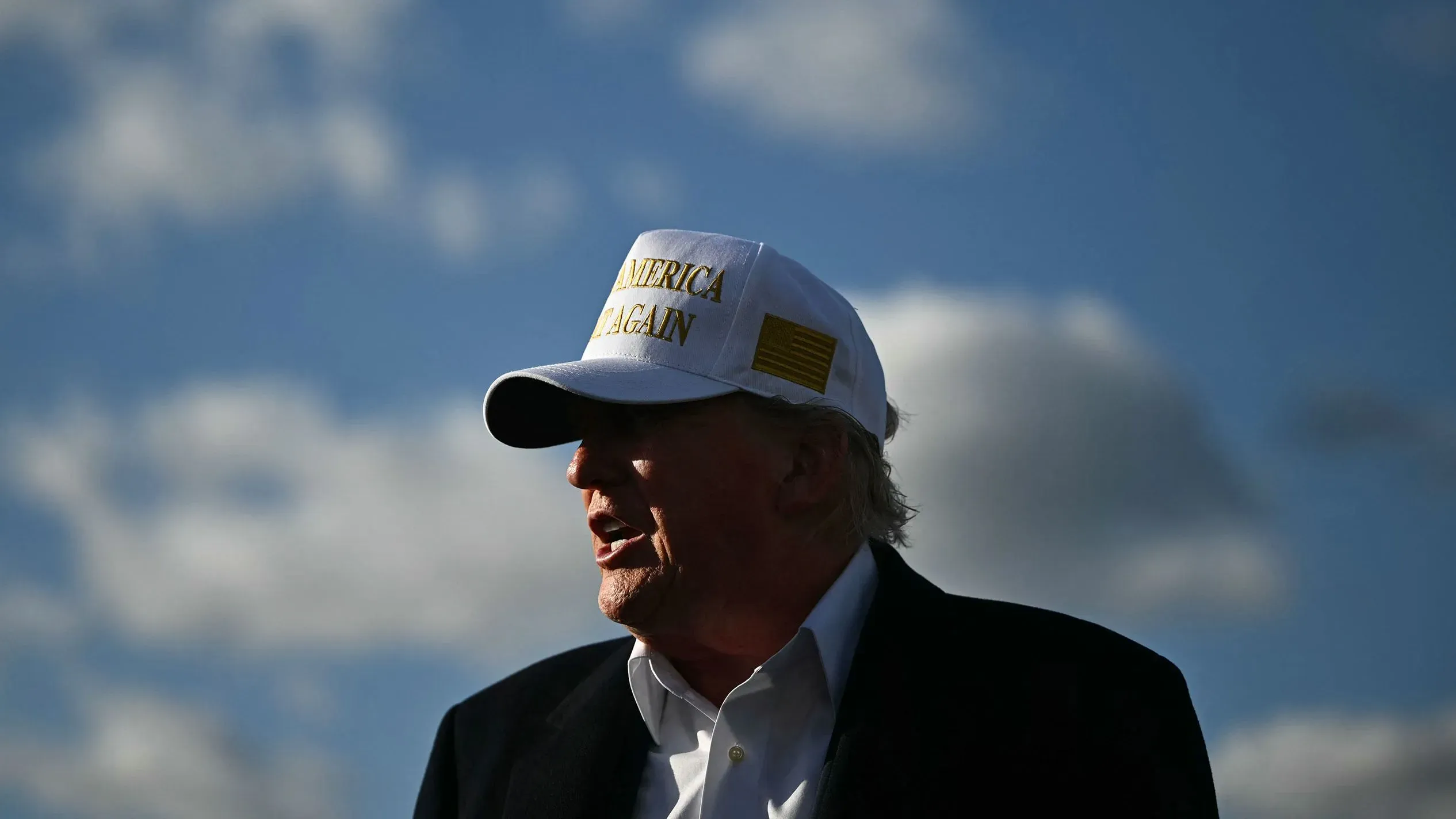 ترامب يتحدث في حدث عام، مرتديًا قبعة مكتوب عليها "اجعل أمريكا عظيمة مرة أخرى"، مع سماء زرقاء وخ Clouds في الخلفية.