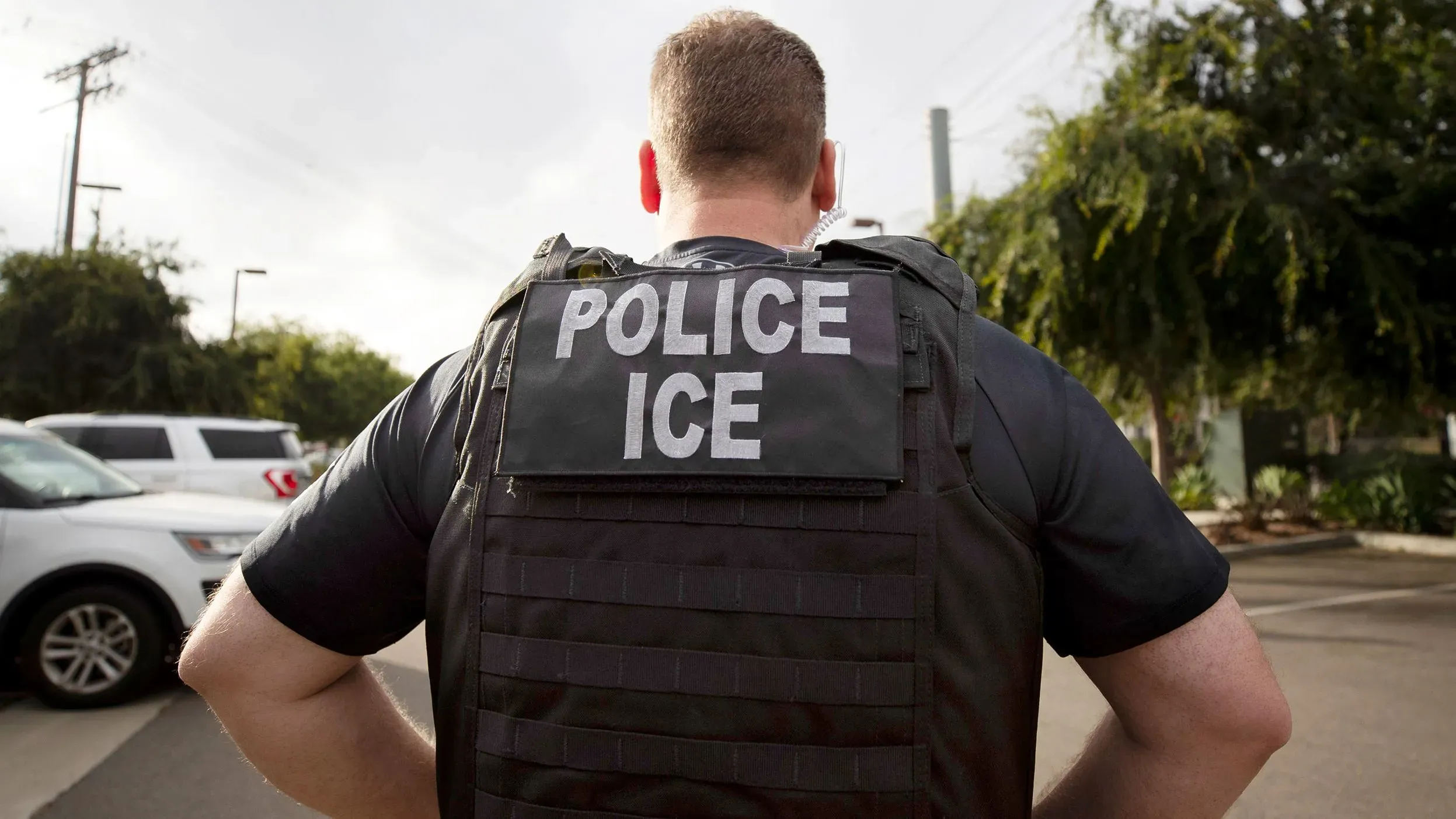 ضابط إنفاذ قانون يرتدي سترة مكتوب عليها \"شرطة ICE\" في موقع خارجي، مما يعكس تغييرات السياسة في اعتقال المهاجرين.